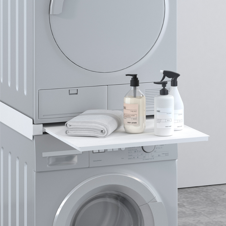 Gray washing machine and dryer stacked, white tray extended with folded towels and laundry supplies. Napofix ML-3 vertical mounting base.