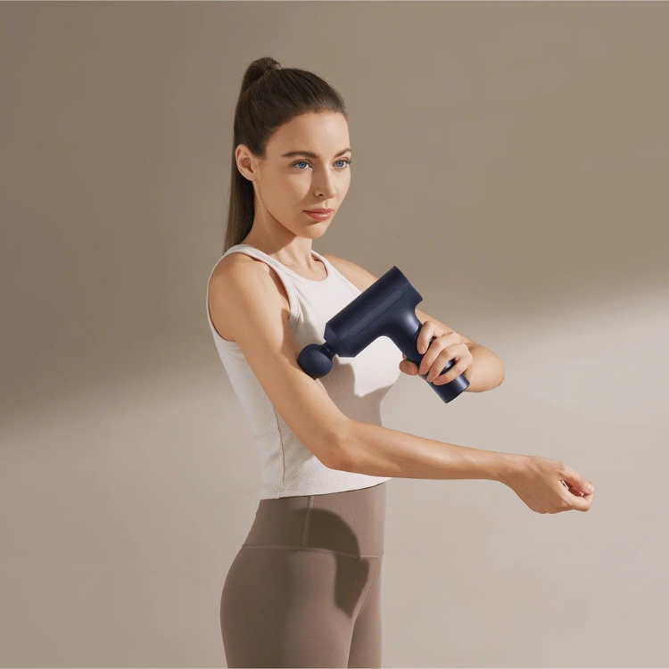 Woman using dark blue Xiaomi massage gun on her arm, wearing white tank top and brown leggings.